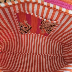 May include: Interior view of a colorful tote bag. The bag features a striped orange and white lining, with a pocket showcasing a tiger print. The exterior has orange, pink, and white stripes.