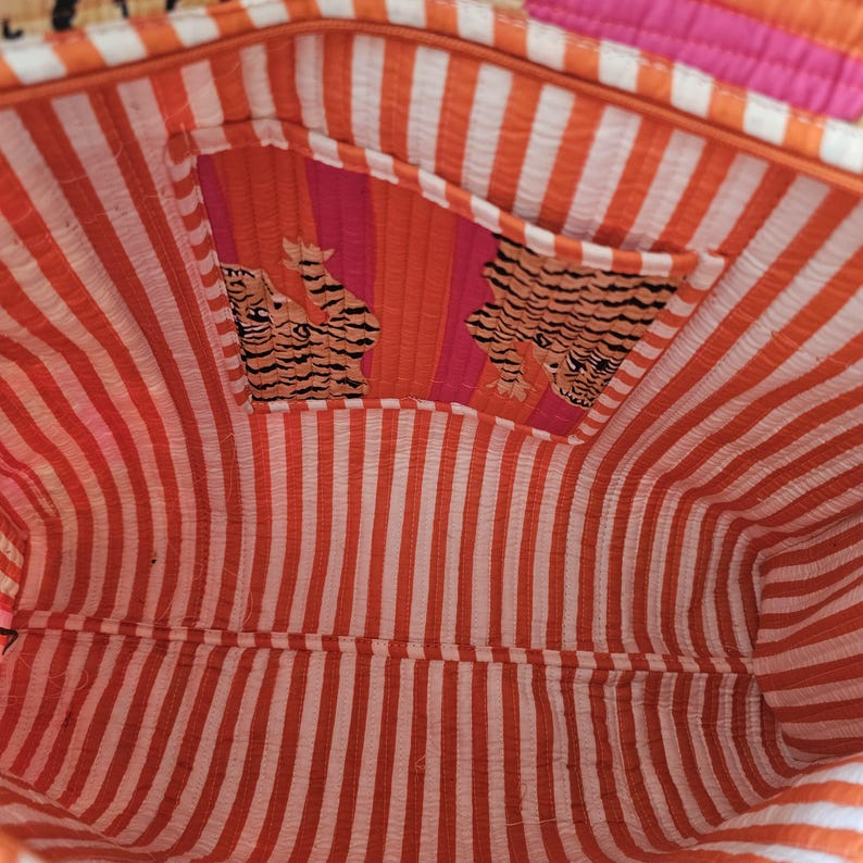 May include: Interior view of a colorful tote bag. The bag features a striped orange and white lining, with a pocket showcasing a tiger print. The exterior has orange, pink, and white stripes.