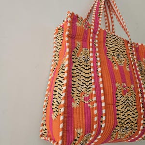 May include: A quilted tote bag with a vibrant tiger print. The bag features orange and pink vertical stripes with tiger illustrations. White and orange striped handles and trim complete the design. The bag is made of a soft, quilted fabric.