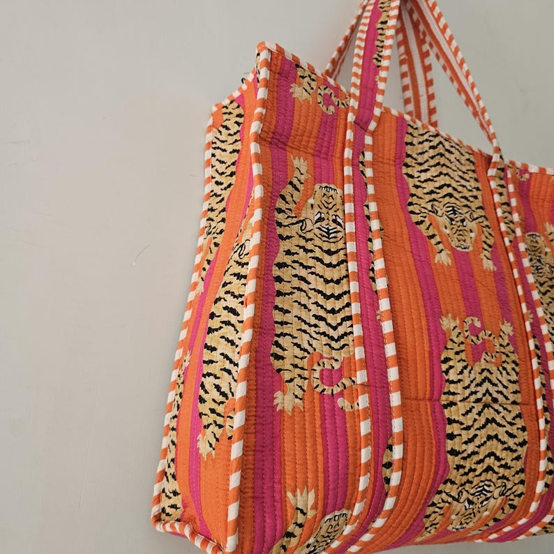 May include: A quilted tote bag with a vibrant tiger print. The bag features orange and pink vertical stripes with tiger illustrations. White and orange striped handles and trim complete the design. The bag is made of a soft, quilted fabric.