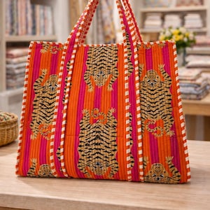 May include: A quilted tote bag with a vibrant tiger print. The bag features a repeating pattern of tigers on a background of orange and pink stripes. The handles are striped with white and orange. The bag is sitting on a wooden table.