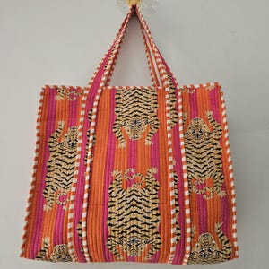 May include: A quilted tote bag with a vibrant tiger print. The bag features a repeating pattern of tigers on a background of vertical stripes in shades of pink and orange. The handles are striped in white and pink.