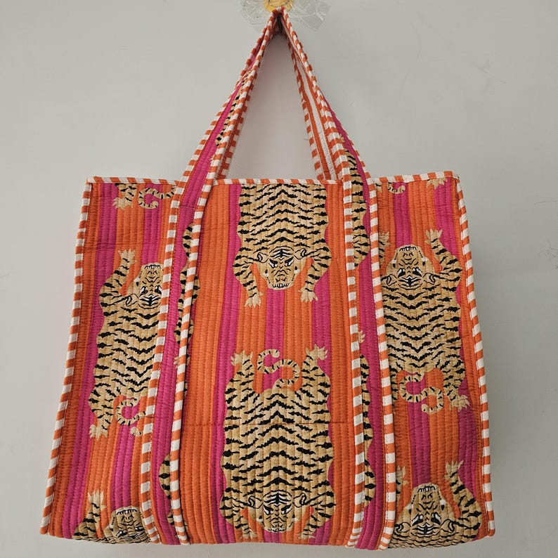 May include: A quilted tote bag with a vibrant tiger print. The bag features a repeating pattern of tigers on a background of vertical stripes in shades of pink and orange. The handles are striped in white and pink.