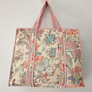May include: Quilted tote bag with a floral and bird pattern in cream, pink, and blue. The bag has pink handles and trim, and a rectangular shape. The fabric has a quilted texture.