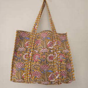 May include: A mustard yellow tote bag with a floral pattern in pink, red, and blue. The bag has long handles and is made of quilted fabric. The floral design is repeated across the entire bag.