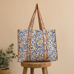 May include: A large tote bag with a floral pattern in blue, pink, and yellow on a white background. The bag has brown handles and trim. The bag is sitting on a small wooden stool.