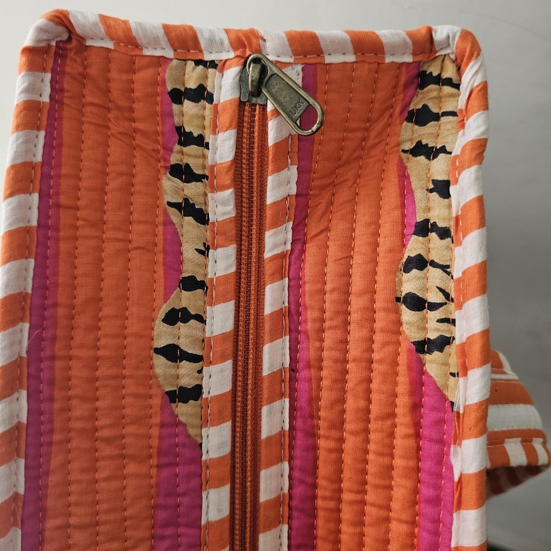May include: Close-up of a quilted fabric with an orange and white striped border. The main body features vertical orange stripes, pink stripes, and decorative patches with black and tan patterns. A bronze zipper runs down the center.
