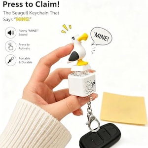 May include: A white seagull keychain featuring a yellow beak and black wing details, mounted on a square white base. The keychain includes a speech bubble graphic with the text MINE! and a metal lobster clasp attachment for portability.