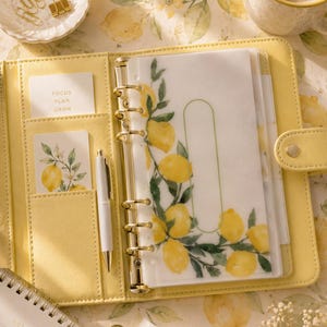 Puede incluir: Un planificador amarillo con detalles dorados, abierto para revelar un diseño de limones. Incluye un bolígrafo blanco, tarjetas con el texto "FOCUS PLAN GROW" y un pequeño bloc de notas. La escena está sobre una superficie con estampado de limones.