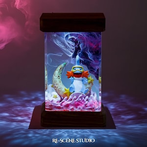 May include: A decorative lamp featuring a blue and white cartoon character with orange accents, wearing goggles, and sitting on a crescent moon. The lamp is encased in a clear rectangular box with a wooden base and top, and the background has a colorful, swirling design.