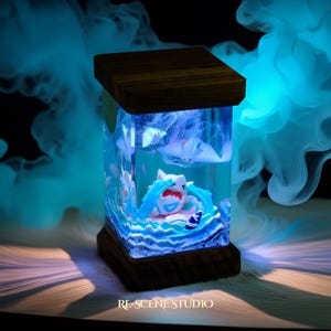 May include: A rectangular, illuminated resin art piece featuring a cartoon character in a watery scene. The piece has a wooden top and base. The character is surrounded by blue waves and other figures. The text "RE-SCENE STUDIO" is at the bottom.