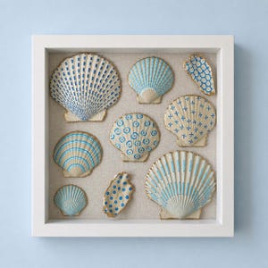 Puede incluir: Un marco cuadrado blanco tipo caja con ocho conchas marinas variadas dispuestas sobre un fondo de tela beige. Cada concha presenta patrones azules pintados a mano, incluyendo rayas, puntos y diseños de cuadrícula, con bordes pintados en dorado.