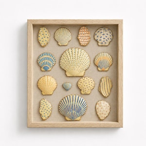 Op de afbeelding: Een ingelijst kunstwerk met een verzameling decoratieve schelpen in verschillende vormen en maten. De schelpen zijn in tinten geel, blauw en oranje, met ingewikkelde patronen en gouden randen. De kunst is geplaatst op een neutrale linnen achtergrond.