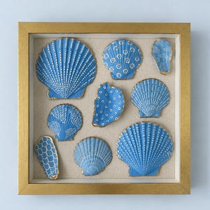 Puede incluir: Un marco cuadrado de tono dorado que contiene ocho conchas marinas azules dispuestas sobre un fondo de tela beige claro. Cada concha presenta patrones pintados en blanco únicos, incluyendo puntos, líneas y escamas, con bordes dorados que resaltan sus formas.