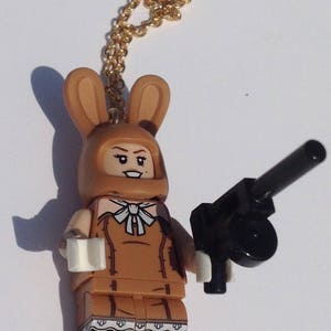 May include: A brown Lego minifigure wearing a bunny costume and holding a black gun. The minifigure has a gold chain necklace.