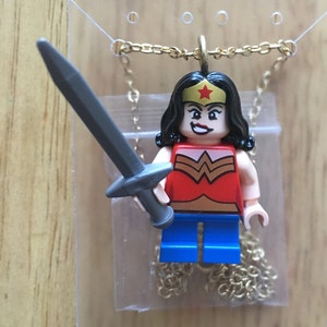 May include: A gold chain necklace featuring a Lego figure of Wonder Woman holding a sword. The figure is wearing a red and gold costume with a blue skirt and a red star on her forehead.