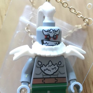 May include: A grey Lego minifigure with a white fur collar and a gold chain necklace. The minifigure has a white beard, red eyes, and a menacing expression. The minifigure is wearing green trousers and a brown belt.
