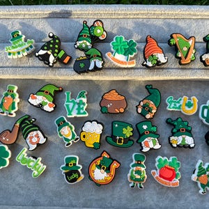 May include: A selection of St. Patrick's Day charms featuring gnomes, shamrocks, pots of gold, beer mugs, and hats. The charms are predominantly green, with orange, black, and white accents. These decorative charms are ideal for shoes or accessories.
