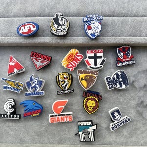 May include: A selection of AFL team logo charms, featuring logos for teams like the Western Bulldogs, Essendon, and others. The charms are in various shapes and colors, with team names and logos displayed.