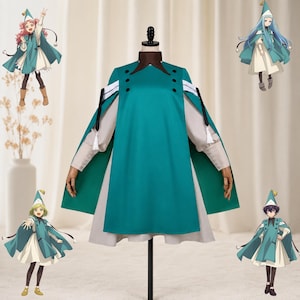 May include: A teal cape-style garment with a brown high collar and black buttons, layered over a light beige long-sleeved dress. The ensemble features white and black ribbon accents at the shoulders and is displayed on a mannequin.