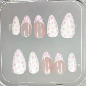 May include: A set of ten oval press-on nails. The nails have a white base with light purple polka dots. Some nails have a nude-coloured tip with a white and light purple polka dot design.