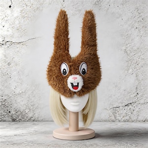 以下が含まれることがあります： 長い耳と漫画の顔が付いた茶色のぬいぐるみウサギの帽子。大きな目、ピンクの鼻、笑顔の口が含まれています。 帽子は、ブロンドの髪のマネキンの頭に表示されています。