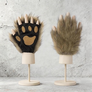 Op de afbeelding: Een paar berenpoot handschoenen. Eén handschoen is zwart met bruine voetzolen en klauwen. De andere handschoen is bruin en harig. Beide handschoenen worden op crèmekleurige standaards getoond.