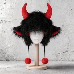 May include: A black faux fur hat with red horns and ear accents. The hat has long black fur and two red pom-poms hanging from black strings. The hat is displayed on a mannequin head.