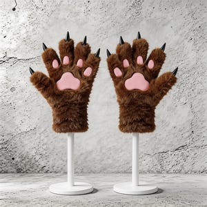 May include: A pair of brown faux fur gloves shaped like bear paws, each with pink paw pads and black claws. The gloves are displayed on white stands against a textured gray background.