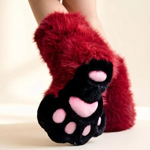 May include: A pair of plush, fuzzy leg warmers or boot covers in a deep burgundy red color. The bottom section features a black faux fur paw design with soft, light pink padded toe beans and a larger central pad, mimicking an animal paw shape.