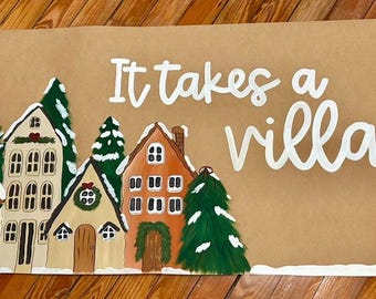 May include: A hand-painted sign on brown paper with the phrase "It takes a village" in white script. The bottom of the sign features a row of colorful houses and evergreen trees, with a snow-covered base. The houses are painted in various colors.