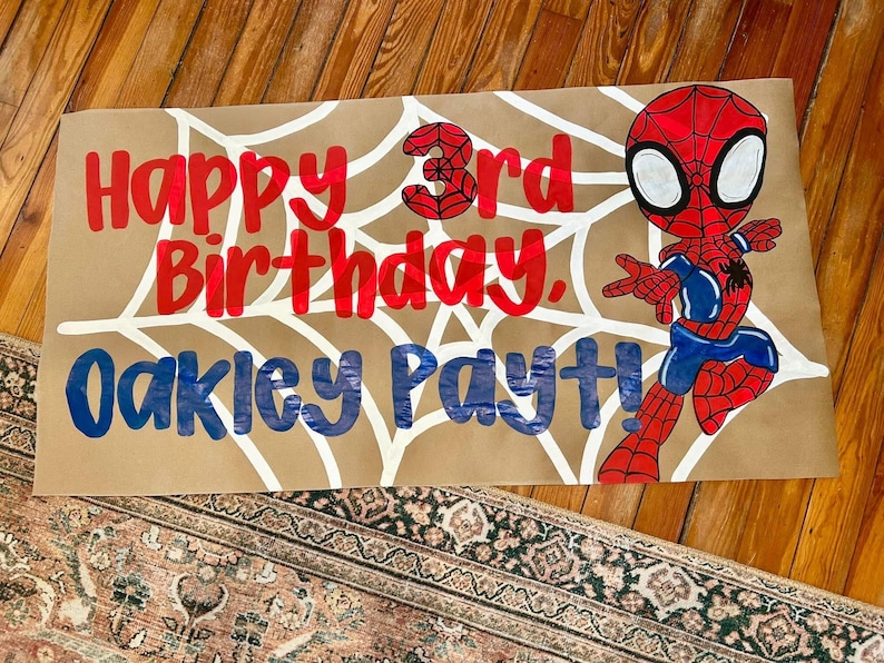 May include: A brown paper banner with a white spiderweb design, red text that reads "Happy 3rd Birthday." and blue text that reads "Oakley Payt!" A cartoon Spider-Man figure is on the right.
