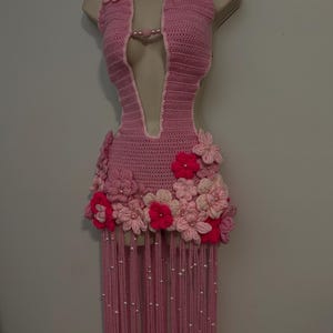 May include: A light pink crochet halter top featuring a deep V-neckline, adorned with various shades of pink and fuchsia crocheted floral appliqués at the neckline and hem, finished with long pink fringe strands embellished with small white beads.