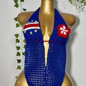 May include: A blue crochet halter top featuring a Cape Verde flag design on the right cup with yellow stars, a white stripe, and a red stripe. The left cup is adorned with a large red and white crocheted flower applique. The garment has a deep V-neck.