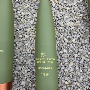 May include: Olive green projectile with a silver tip and copper band. The text "155 TNT PROJECTILE M795 W/SUPPL CHG" is printed on the side, along with other markings. The projectile is cylindrical with a tapered nose.