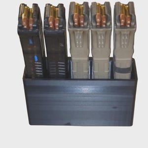 May include: A black plastic magazine holder containing four loaded magazines. The magazines are black and tan with visible brass-colored bullets. The holder is rectangular and sits on a light gray background.
