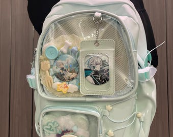 Puede incluir: Una mochila azul claro con múltiples compartimentos y bolsillos transparentes. La mochila presenta pines decorativos, dijes y un accesorio esponjoso. El diseño incluye detalles de estrellas y nubes, y un pequeño tarjetero.