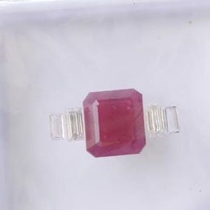 Puede incluir: Un anillo con una gran piedra preciosa rectangular de color rosa intenso en el centro, flanqueada a cada lado por dos piedras más pequeñas, transparentes y rectangulares de corte baguette. El anillo se presenta en un joyero cuadrado de plástico blanco.