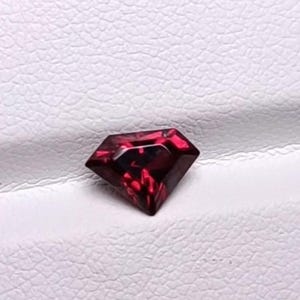 Könnte beinhalten: Ein facettierter, tiefroter Edelstein im Brillantschliff mit scharfen geometrischen Kanten und einer reflektierenden Oberfläche. Der Stein ist auf einem strukturierten, weißen, gefalteten Material platziert, das einen Kontrast zum Farbton bildet.