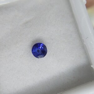 Könnte beinhalten: Ein runder, facettierter, tiefblauer Edelstein. Der Stein ist vor einem weißen Hintergrund platziert, der seinen Brillantschliff und seine Farbe hervorhebt. Dieser Edelstein ist wahrscheinlich ein Saphir, perfekt für Schmuck.
