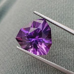 Könnte beinhalten: Ein facettierter, schildförmiger violetter Edelstein, der von einer silberfarbenen Metallpinzette gehalten wird. Der Stein weist einen komplexen, symmetrischen geometrischen Schliff auf, der tiefe violette und helllavendelfarbene Reflexe erzeugt.
