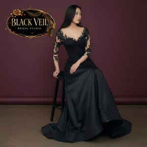 Puede incluir: Vestido de novia negro con escote en V y mangas de encaje. El vestido presenta una falda fluida y es de Black Veil Bridal Studio. La imagen muestra a una mujer sentada en un taburete, mostrando el diseño y el ajuste del vestido.