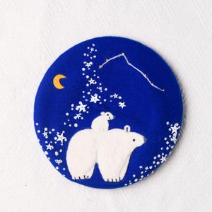 May include: A round, blue beret featuring a whimsical design of two white polar bears under a starry night sky. A small moon and a constellation are also depicted. The beret is a stylish accessory.
