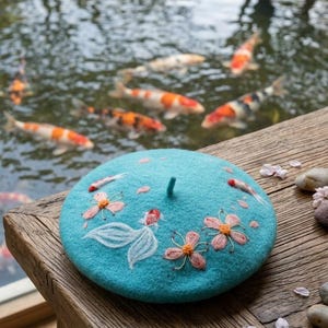 Puede incluir: Un boina turquesa con bordados de peces koi y flores de cerezo. La boina es de fieltro y está sobre una superficie de madera. Peces koi nadan en el fondo.