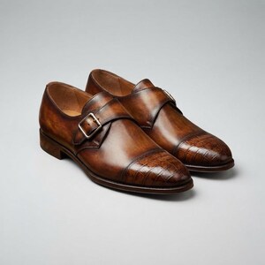 May include: A pair of brown leather monk strap shoes featuring a polished finish with a burnished gradient effect. The shoes have a single silver-tone metal buckle closure and a textured crocodile-embossed pattern on the rounded toe cap.