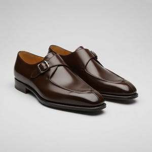 Op de afbeelding: Een paar donkerbruine leren schoenen met een enkele gespsluiting en een gepolijste afwerking, voorzien van een zilverkleurige metalen gesp aan de zijkant, een ronde neus, een lage hak en een gladde leren bovenkant.