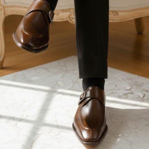 Op de afbeelding: Een paar bruine leren schoenen met een enkele zilverkleurige gespsluiting, gedragen met zwarte sokken en een zwarte broek. De schoenen hebben een gladde afwerking, een afgeronde neus en een opvallende gestikte naad over de wreef.