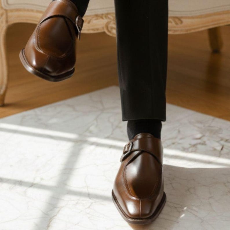 Op de afbeelding: Een paar bruine leren schoenen met een enkele zilverkleurige gespsluiting, gedragen met zwarte sokken en een zwarte broek. De schoenen hebben een gladde afwerking, een afgeronde neus en een opvallende gestikte naad over de wreef.
