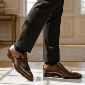Op de afbeelding: Een paar gepolijste bruine leren monkstrap schoenen met een enkele zilverkleurige metalen gespsluiting over de wreef, een ronde neus en een lage hak, gedragen met een donkere broek en zwarte sokken.