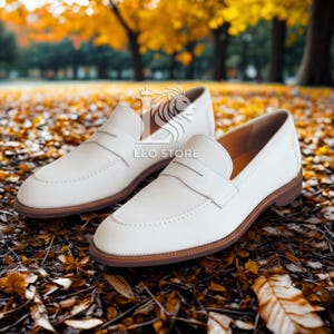 Op de afbeelding: Een paar witte leren loafers met bruine zolen. De schoenen hebben een klassiek ontwerp met een instapmodel en een band over de bovenkant. De afbeelding is buiten gemaakt met herfstbladeren op de achtergrond.